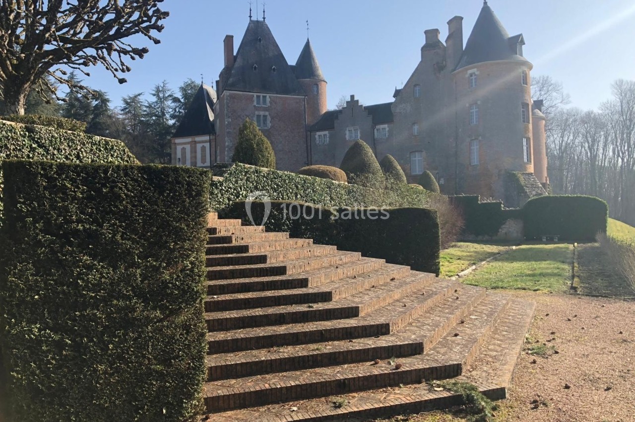 Escalier en pierre menant à un château en briques entouré de haies taillées et d'arbres sous un ciel dégagé.