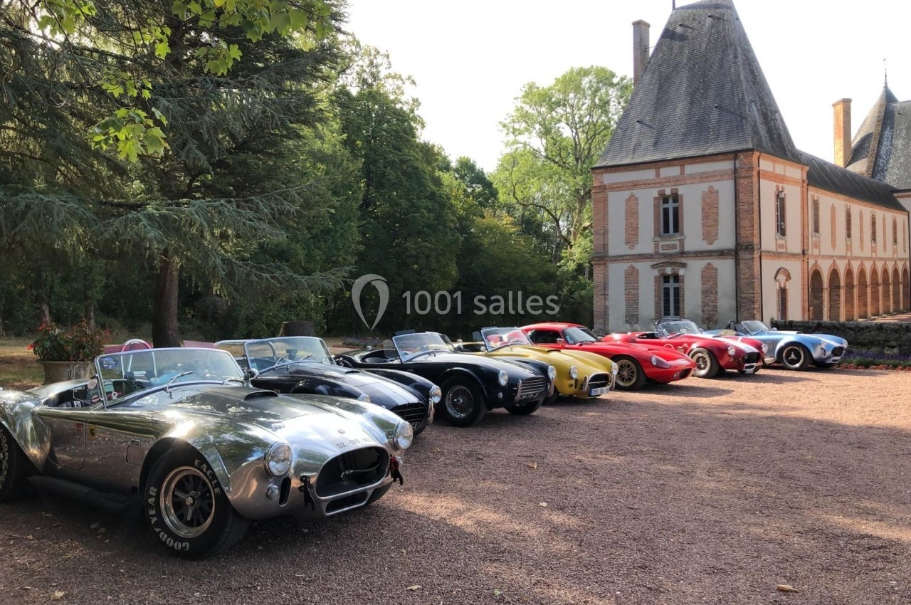 Alignement de voitures de sport classiques devant un bâtiment historique entouré de verdure.