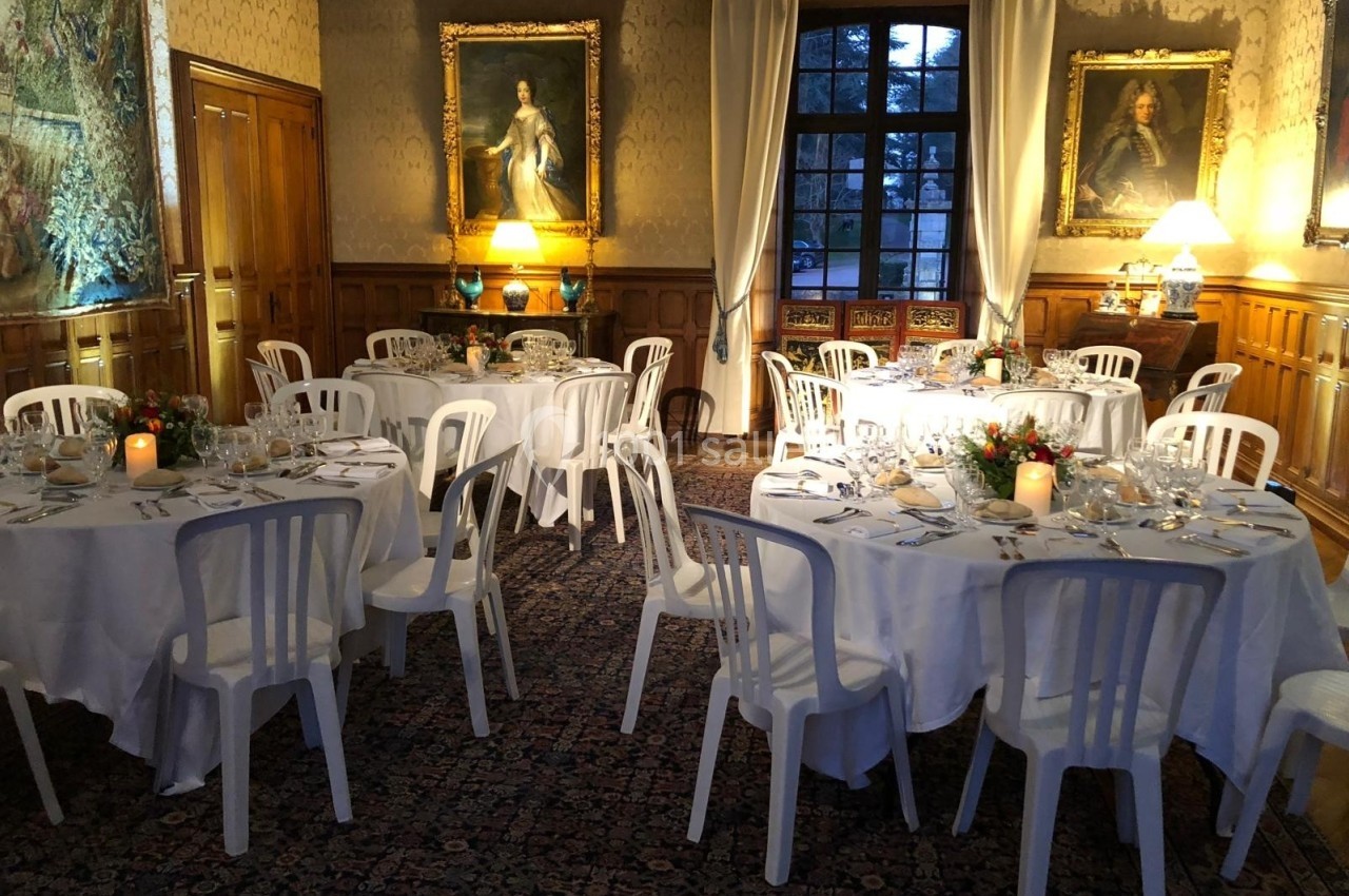 Salle élégante avec tables rondes dressées, chaises blanches, tableaux anciens, tapis et lumière tamisée.