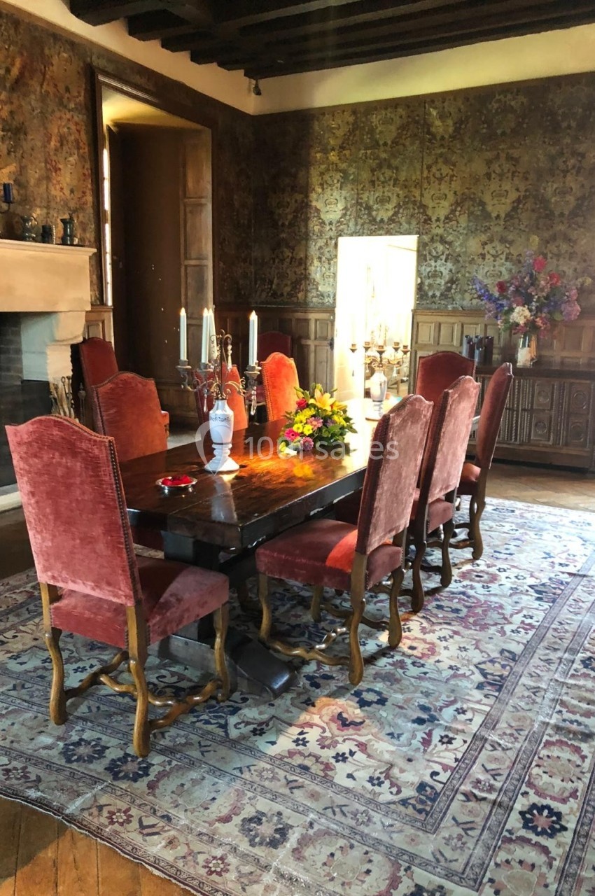 Salle à manger avec table en bois, chaises rouges, tapis persan, cheminée et décoration florale.