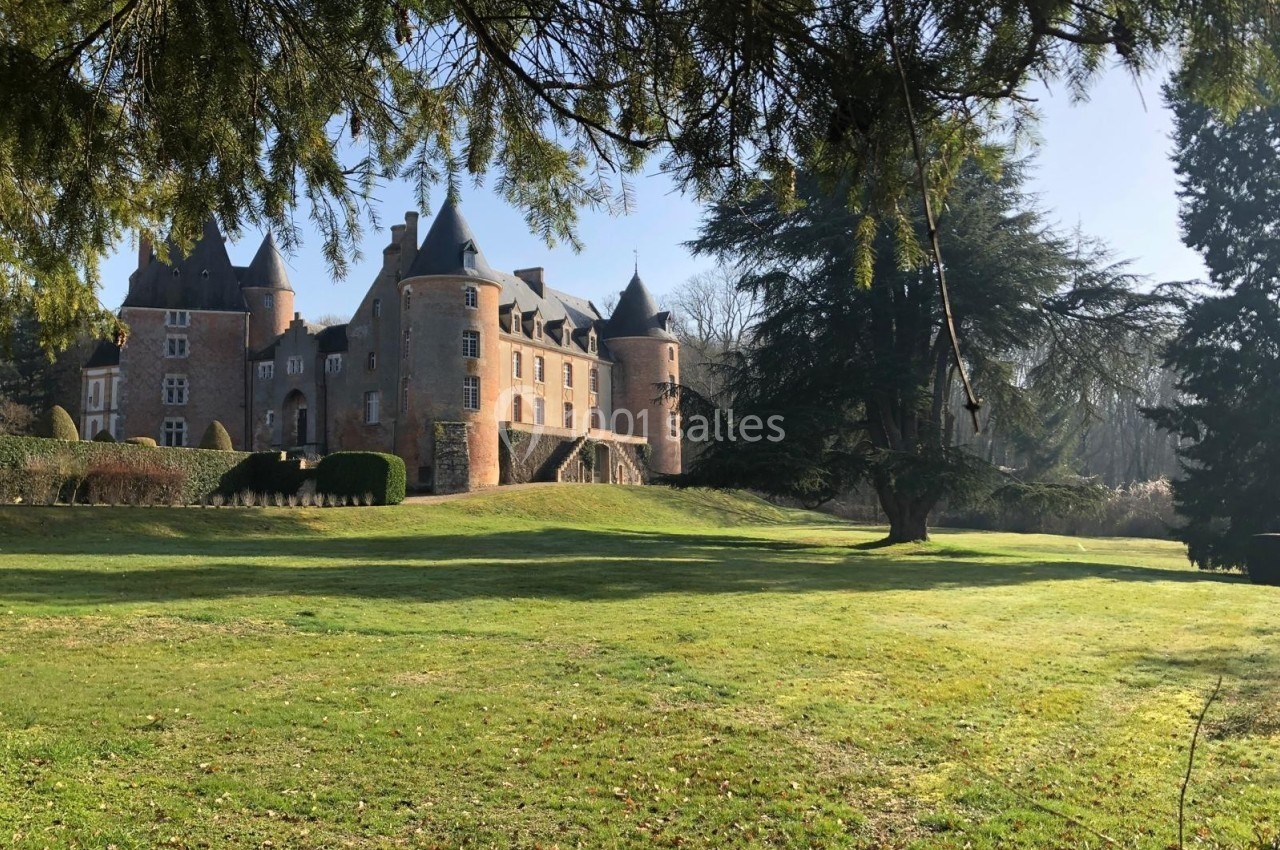 Château en briques entouré d'un grand parc verdoyant, sous un ciel clair et encadré par des arbres.