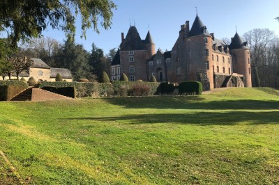 Château en briques rouges entouré de pelouses et d'arbres, sous un ciel clair et ensoleillé.