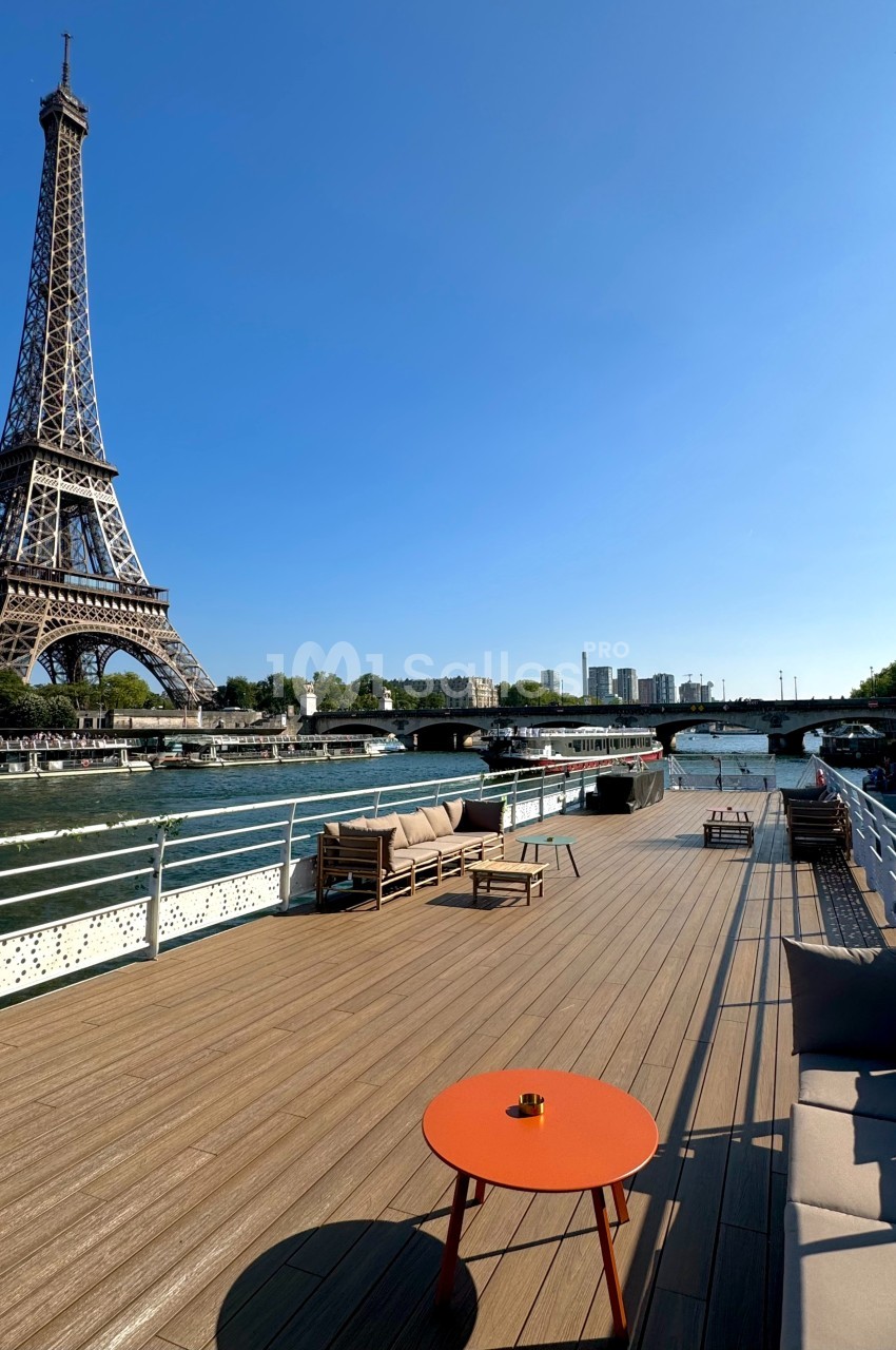 Vue d'une terrasse en bois sur une péniche, avec la tour Eiffel et la Seine en arrière-plan par temps ensoleillé.