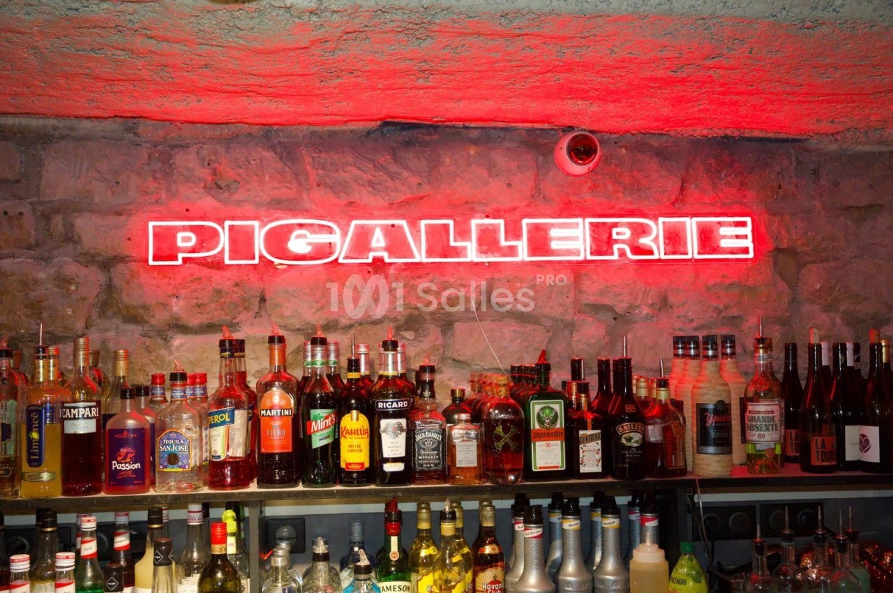 Bar avec un mur en pierre, des bouteilles d'alcool alignées et une enseigne lumineuse rouge indiquant ’Pigallerie’.