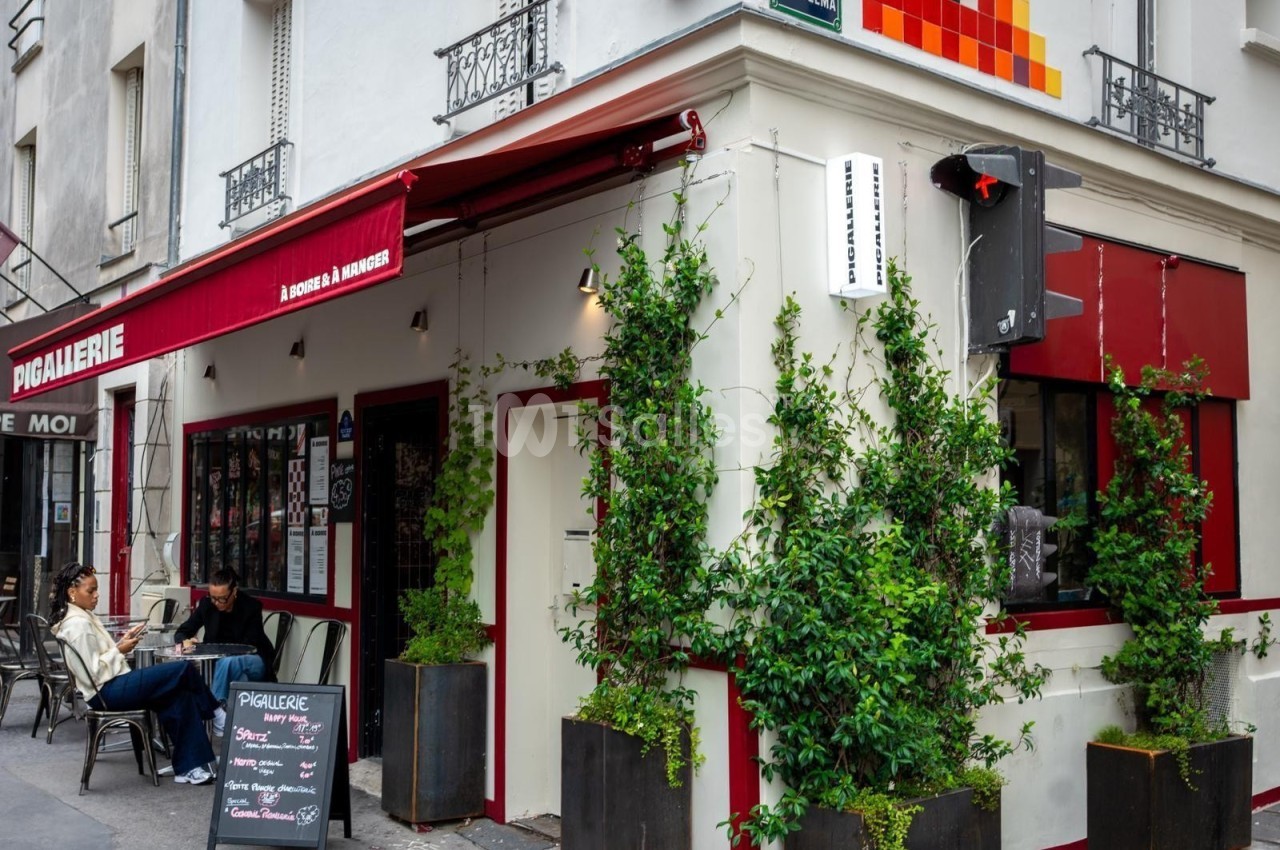 Façade d'un café avec terrasse, plantes grimpantes et enseigne rouge, situé à un coin de rue en ville.