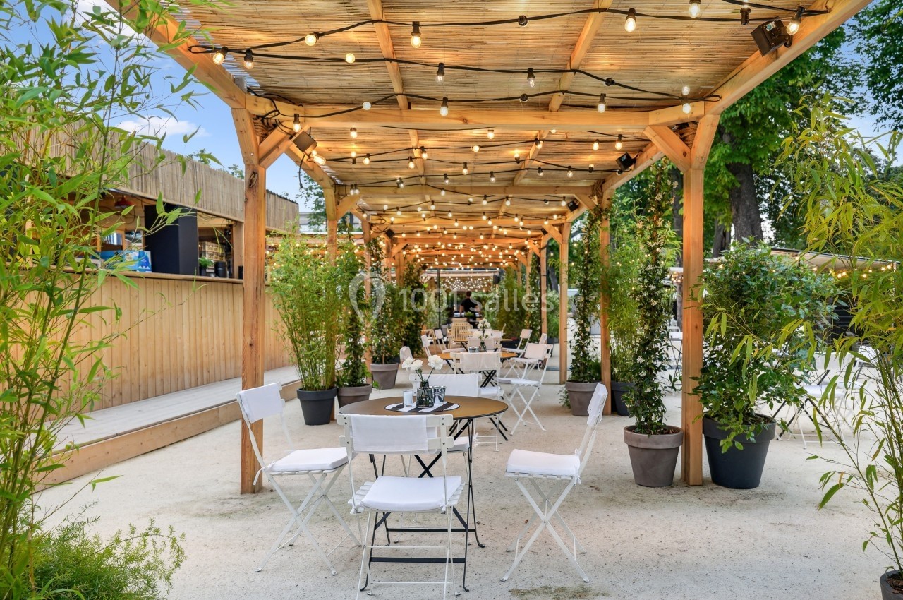 Terrasse extérieure avec pergola en bois, guirlandes lumineuses, tables et chaises blanches entourées de plantes.