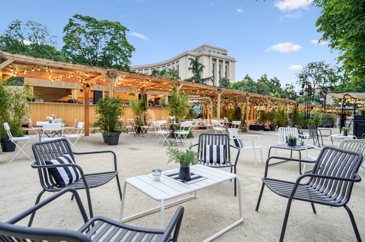 Terrasse extérieure avec tables et chaises, entourée de végétation, éclairée par des guirlandes lumineuses.
