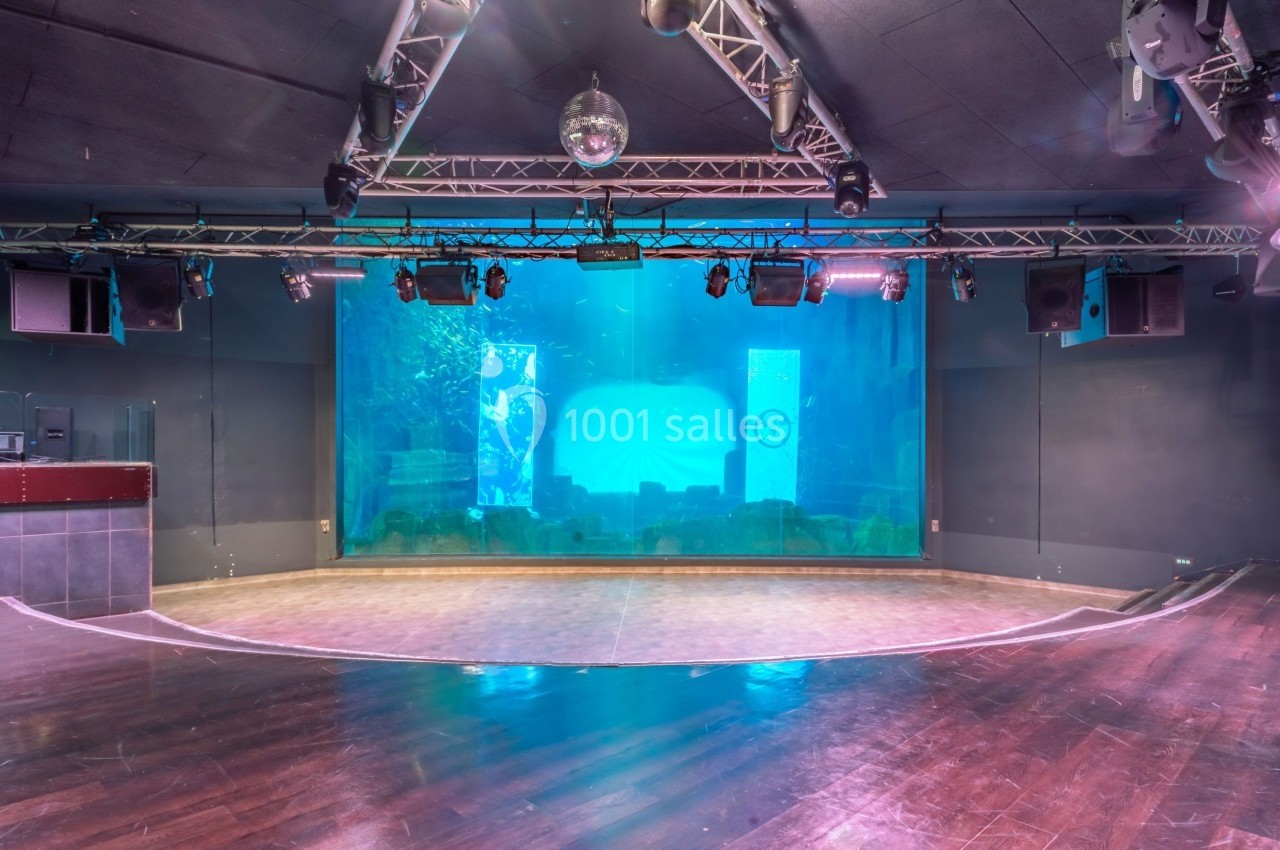 Scène vide avec un grand aquarium en arrière-plan, éclairée par des projecteurs suspendus au plafond.
