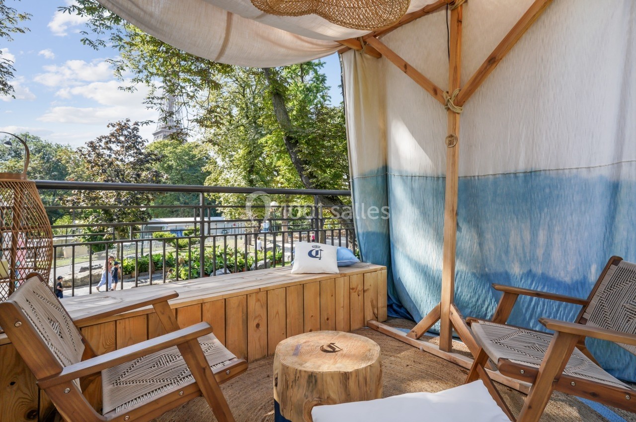 Espace extérieur avec deux chaises en bois, une table basse et un banc, entouré de verdure sous une toile ombragée.