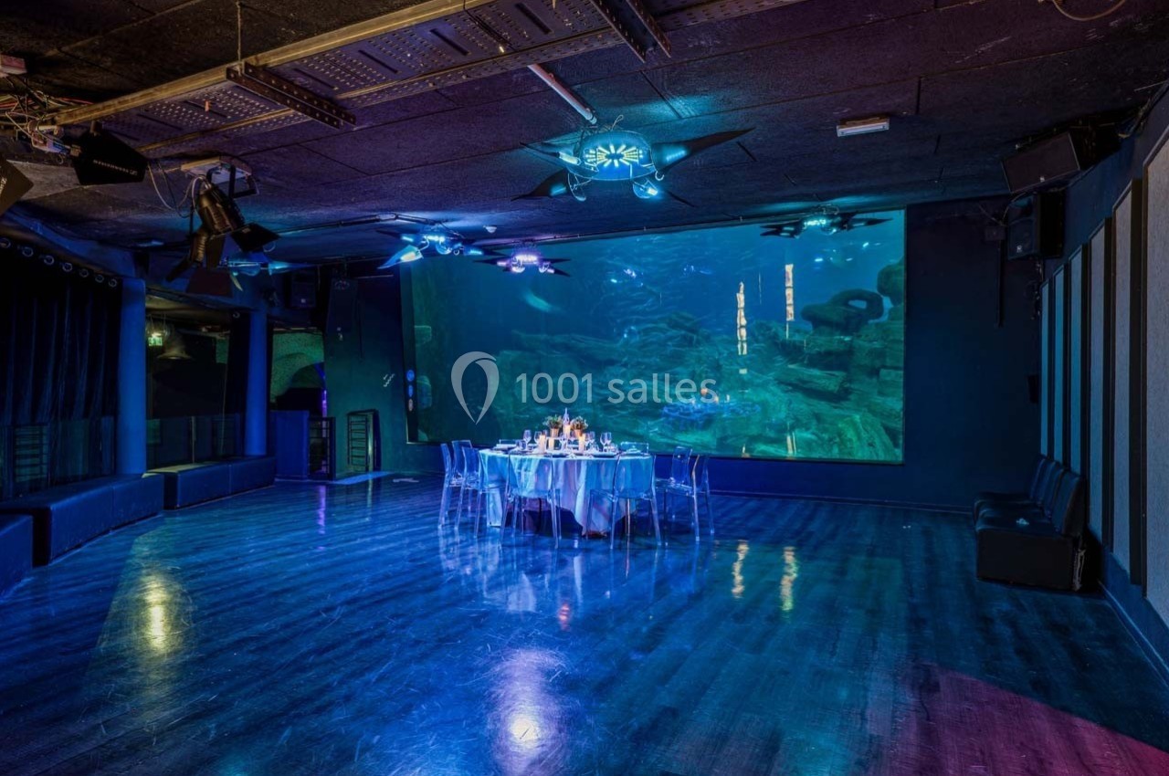 Salle sombre avec un écran géant affichant un décor sous-marin, table dressée au centre et éclairages bleutés.