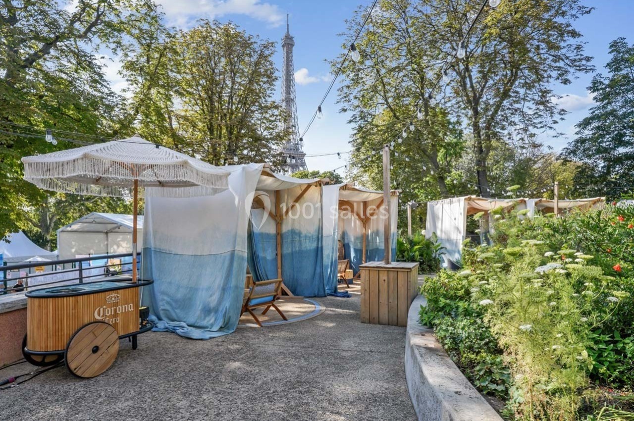 Terrasse aménagée avec chaises longues et parasols, vue sur la tour Eiffel à travers des arbres et des rideaux légers.