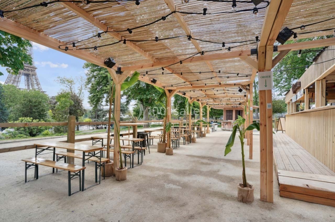 Terrasse extérieure avec tables et bancs en bois sous une pergola, vue sur la Tour Eiffel en arrière-plan.