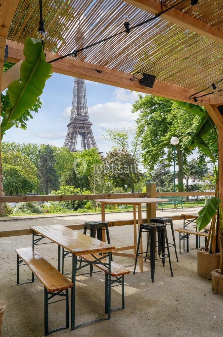 Terrasse en bois avec tables et tabourets, vue sur la tour Eiffel entourée d'arbres par une journée ensoleillée.