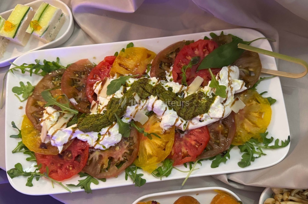 Assiette de tomates colorées avec burrata, pesto, roquette et copeaux de fromage, présentée sur une nappe beige.