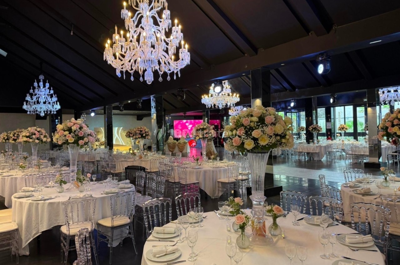 Salle de réception élégante avec tables dressées, chaises transparentes, grands bouquets floraux et lustres en cristal.