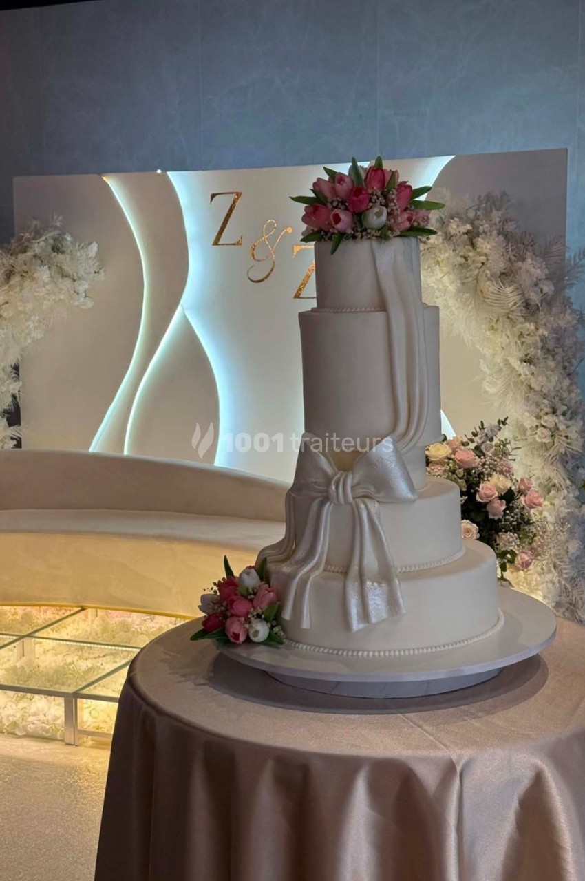 Gâteau de mariage à quatre étages décoré de fleurs roses et d'un nœud blanc, posé sur une table drapée.
