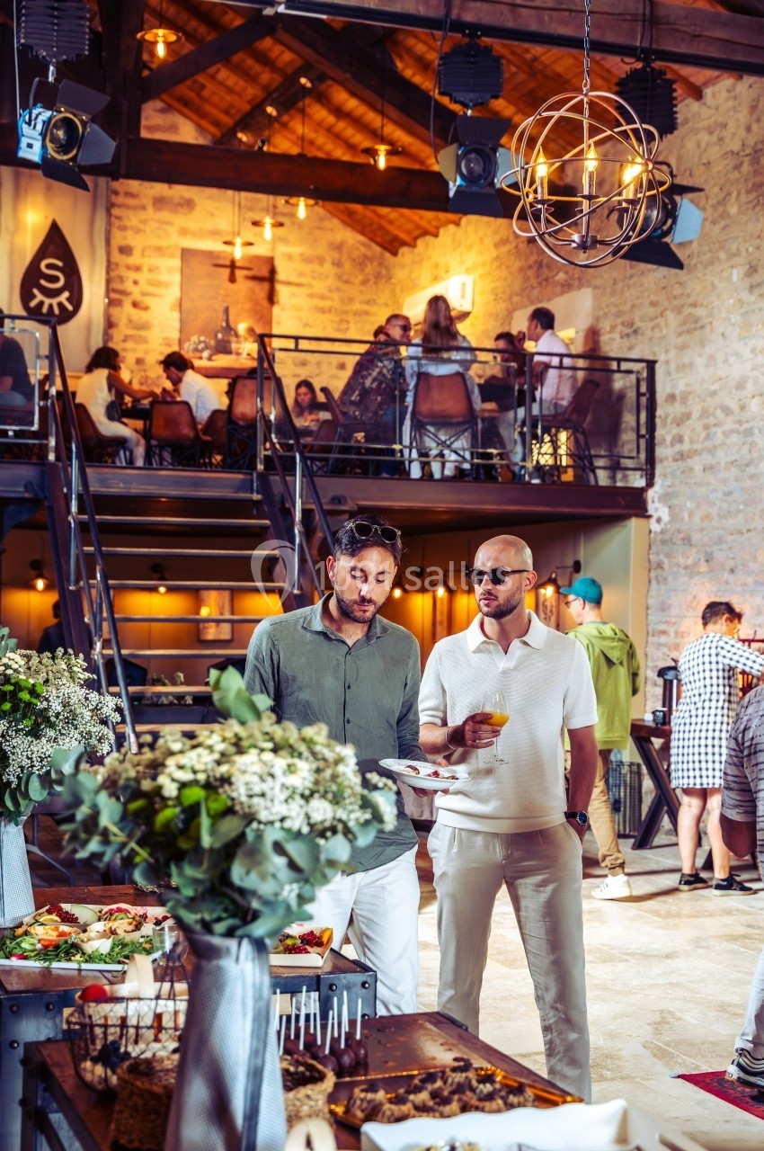Deux hommes discutent près d'un buffet dans une salle avec mezzanine, éclairée par des lumières suspendues.