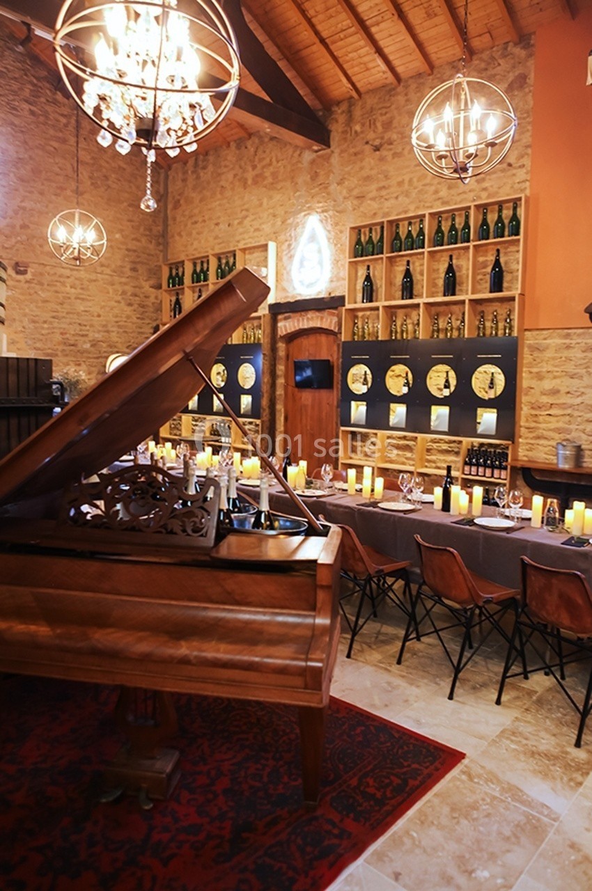 Salle chaleureuse avec murs en pierre, piano à queue, table dressée, chandelles allumées et étagères de bouteilles.