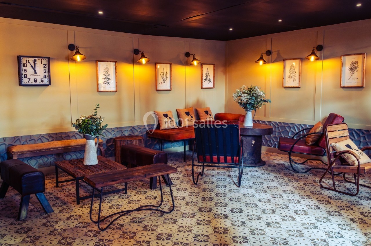 Salon cosy avec mobilier vintage, tables en bois, fauteuils en cuir, décorations murales et éclairage chaleureux.