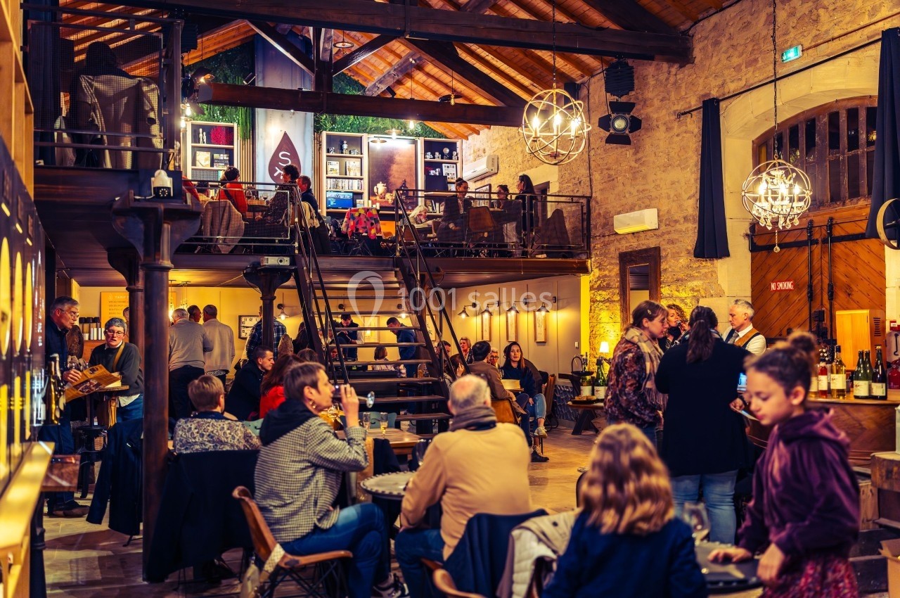 Salle animée avec des tables occupées, un escalier central, des lumières chaleureuses et des murs en pierre apparente.