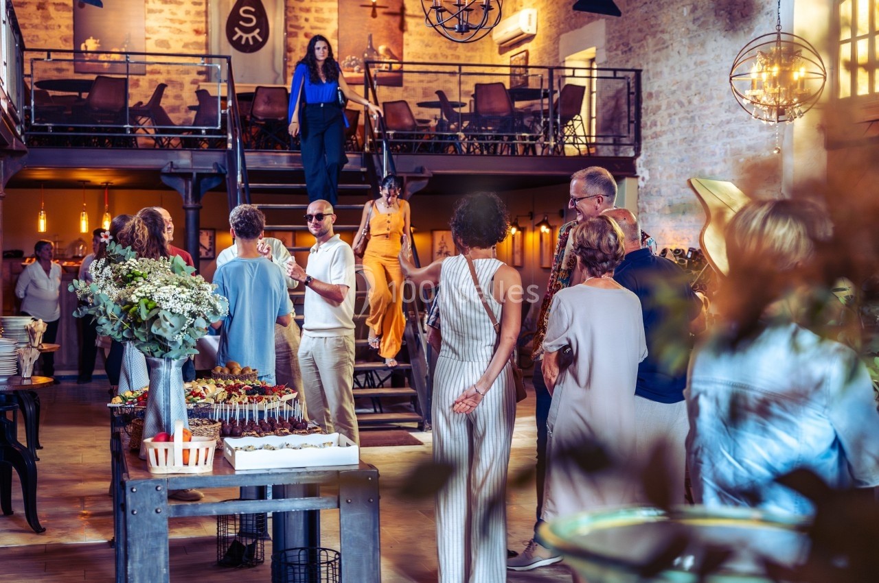 Groupe de personnes discutant dans une salle lumineuse avec mezzanine, décorée de tables et de fleurs.