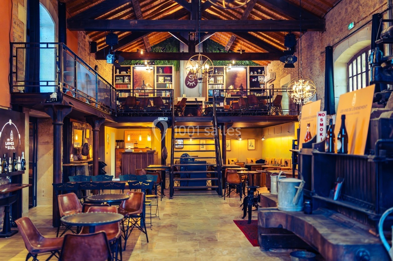 Intérieur d'un bar chaleureux avec mezzanine, tables en bois, étagères de bouteilles et éclairage tamisé.