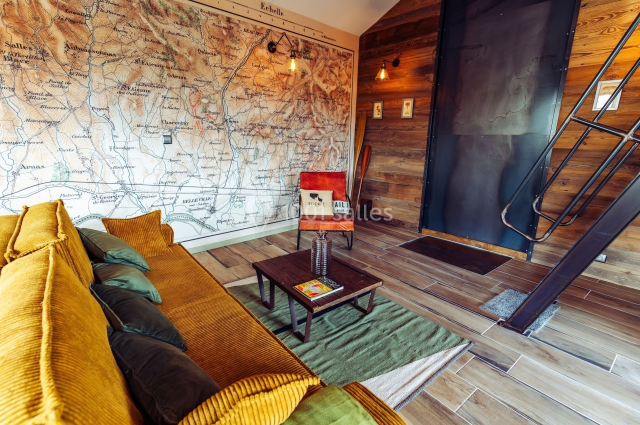 Salon avec canapé jaune, mur décoré d'une carte ancienne, chaise en cuir, table basse et escalier métallique.