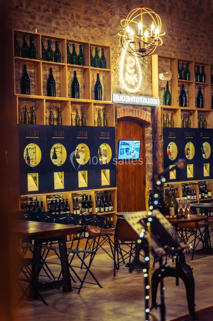 Intérieur d'une cave à vin avec étagères remplies de bouteilles, tables hautes et éclairage chaleureux.