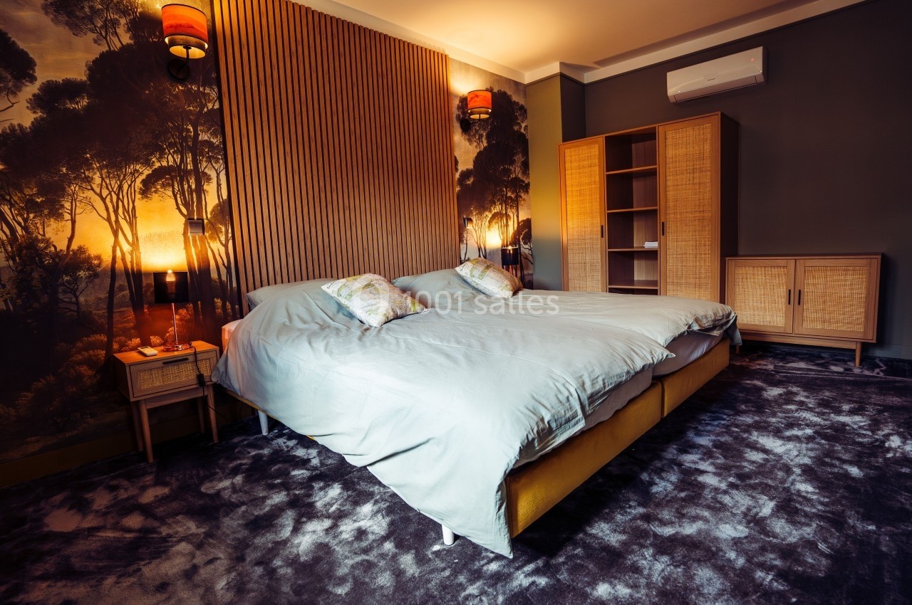 Chambre avec lit double, mur en bois, papier peint nature, meubles en rotin et éclairage tamisé.