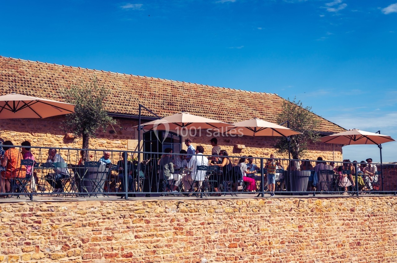 Terrasse ensoleillée avec des tables, des parasols et des personnes attablées devant un bâtiment en pierre.