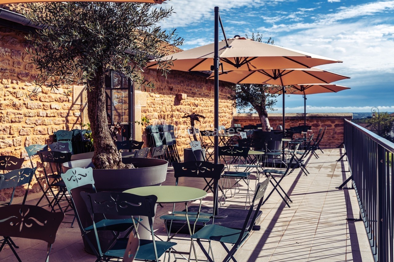 Terrasse ensoleillée avec tables, chaises en métal et parasols, bordée d'un mur en pierre et offrant une vue dégagée.