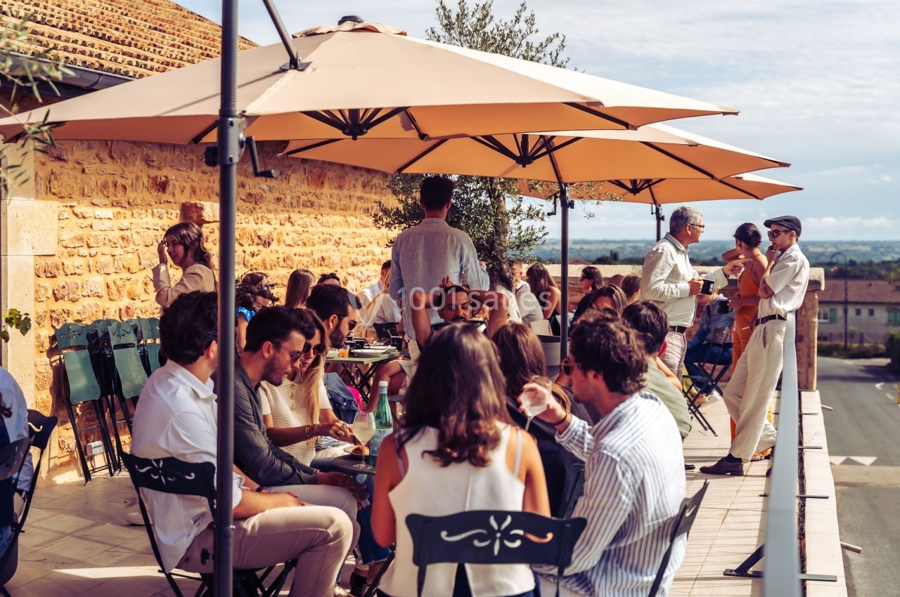Groupe de personnes assises et debout sur une terrasse ensoleillée, discutant sous de grands parasols beige.
