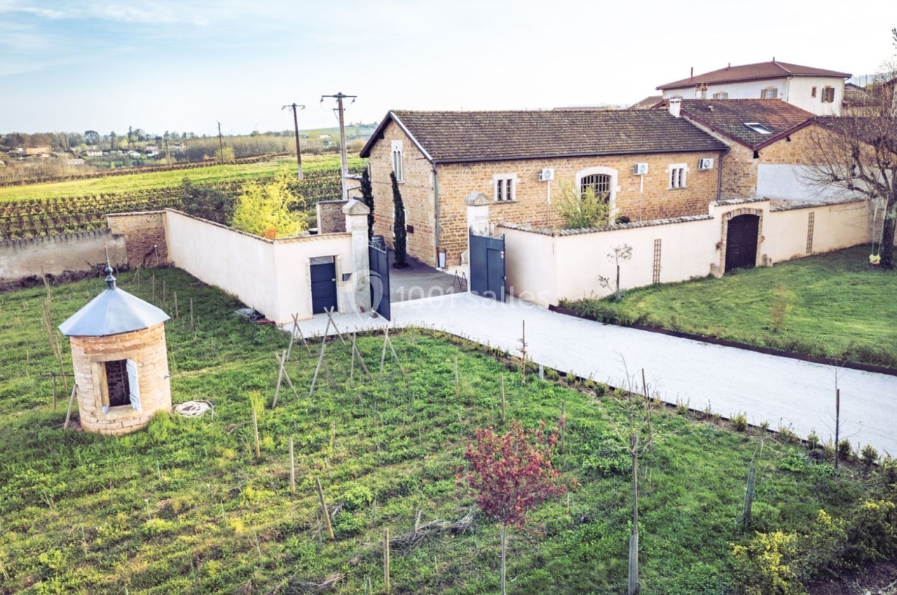 Vue d'un domaine rural avec bâtiments en pierre, allée pavée, petit puits et vignobles en arrière-plan.
