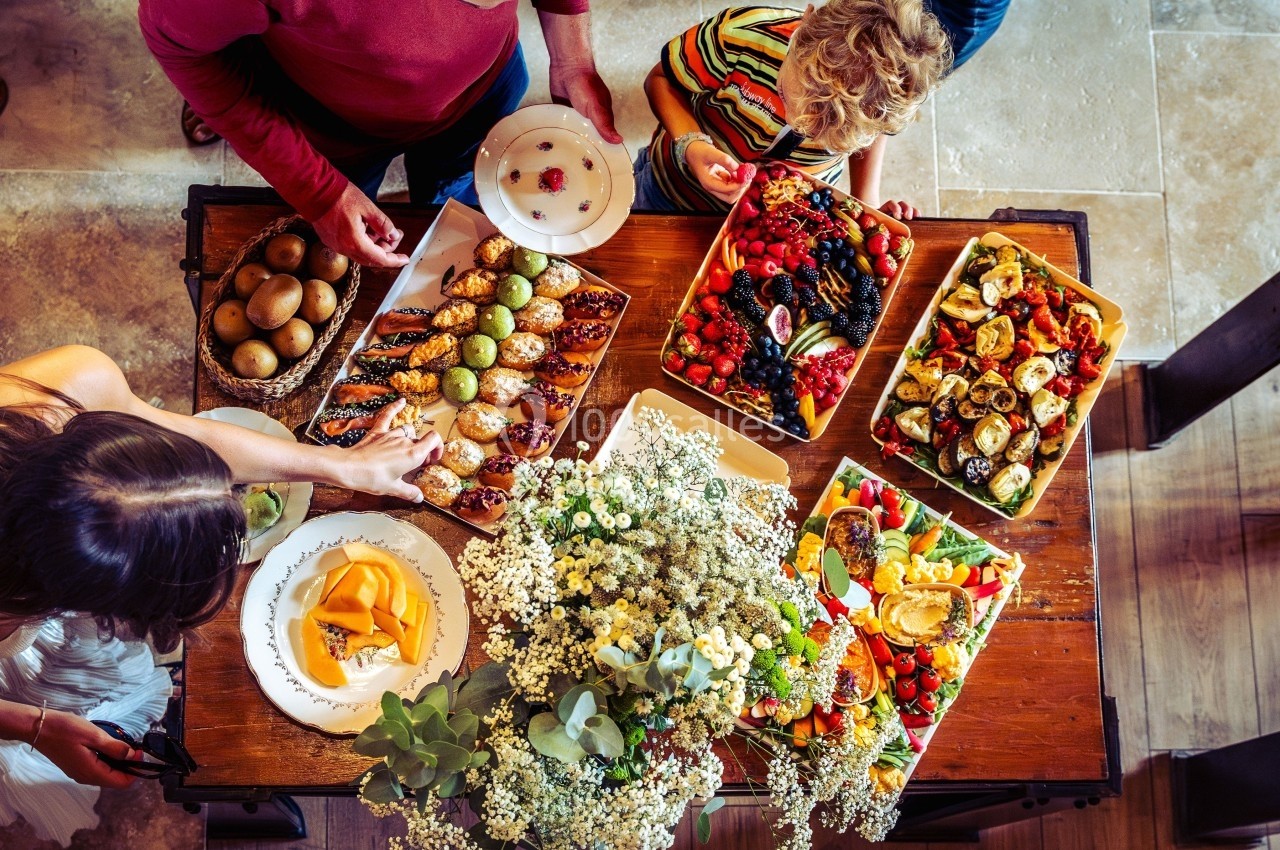 Table vue de dessus avec divers plats colorés, fruits, pâtisseries et fleurs, entourée de personnes se servant.