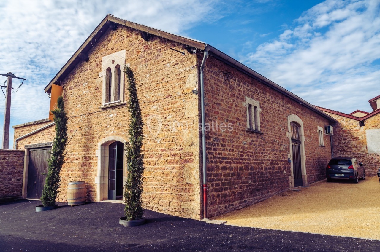 Bâtiment en pierre avec des fenêtres arquées, deux cyprès devant et une voiture garée sur le côté.