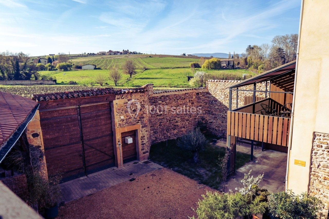 Cour intérieure en pierre avec portail en bois, terrasse couverte et vue sur un paysage rural verdoyant.