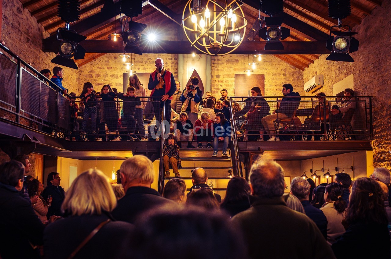 Un groupe de personnes assiste à un discours dans une salle en pierre avec mezzanine et éclairage chaleureux.
