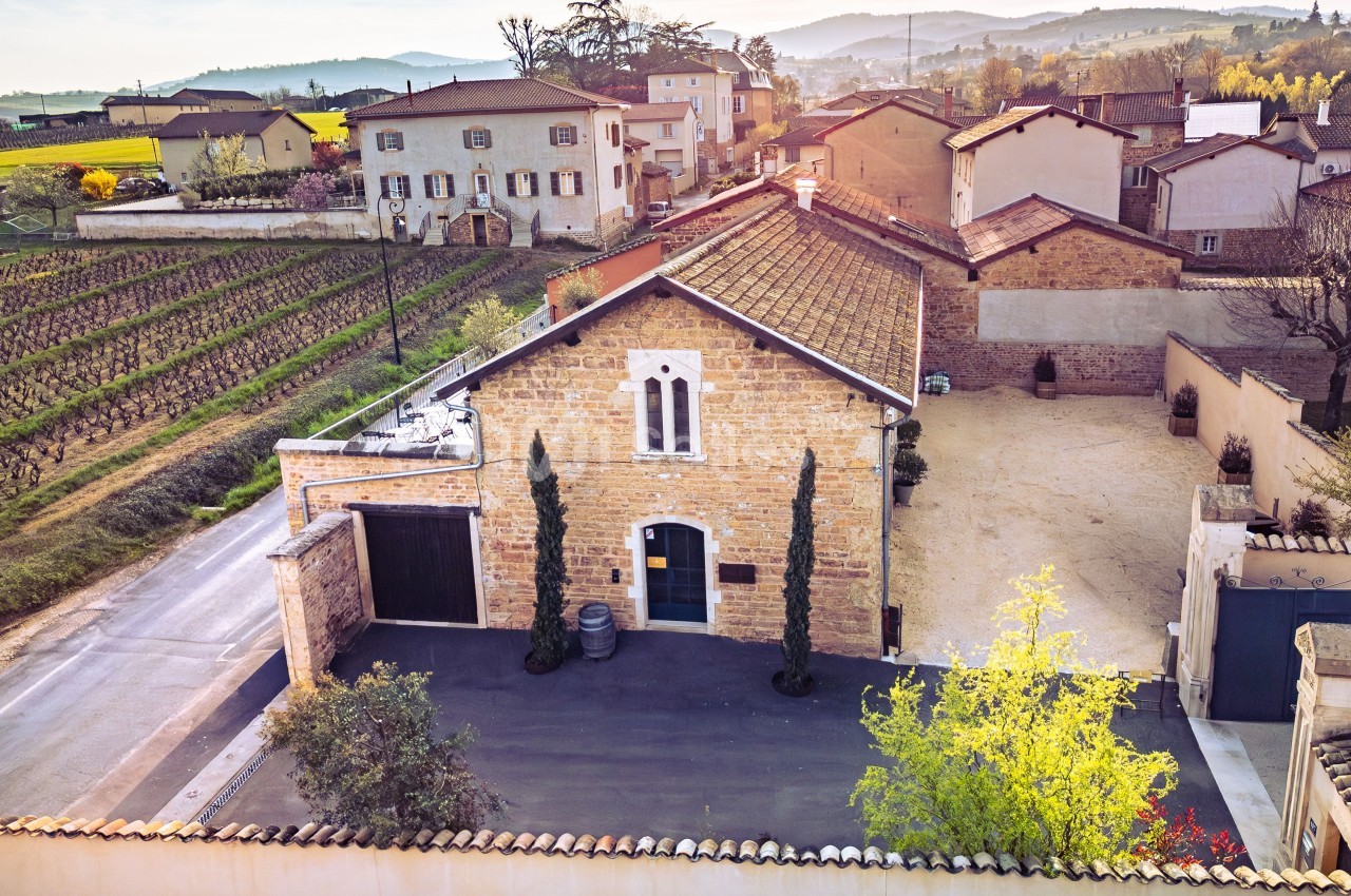 Vue aérienne d'une maison en pierre avec cour, entourée de vignes et de bâtiments dans un paysage rural ensoleillé.