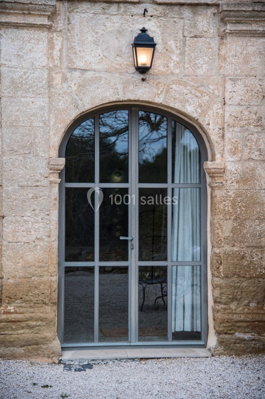 Porte vitrée en arc encadrée de pierre, avec un rideau blanc à l'intérieur et une lampe extérieure allumée au-dessus.