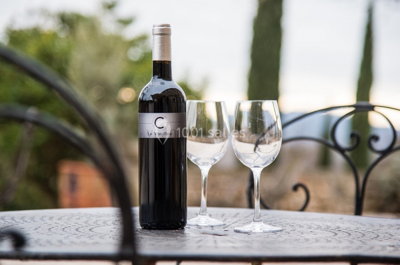 Bouteille de vin rouge et deux verres vides posés sur une table en métal, avec un paysage flou en arrière-plan.