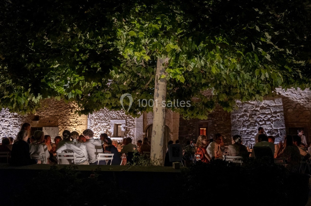 Groupe de personnes dînant en plein air sous un arbre, devant un bâtiment en pierre éclairé la nuit.