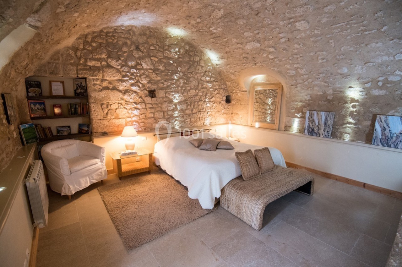 Chambre voûtée en pierre avec lit double, fauteuil, étagère, éclairage chaleureux et décoration minimaliste.