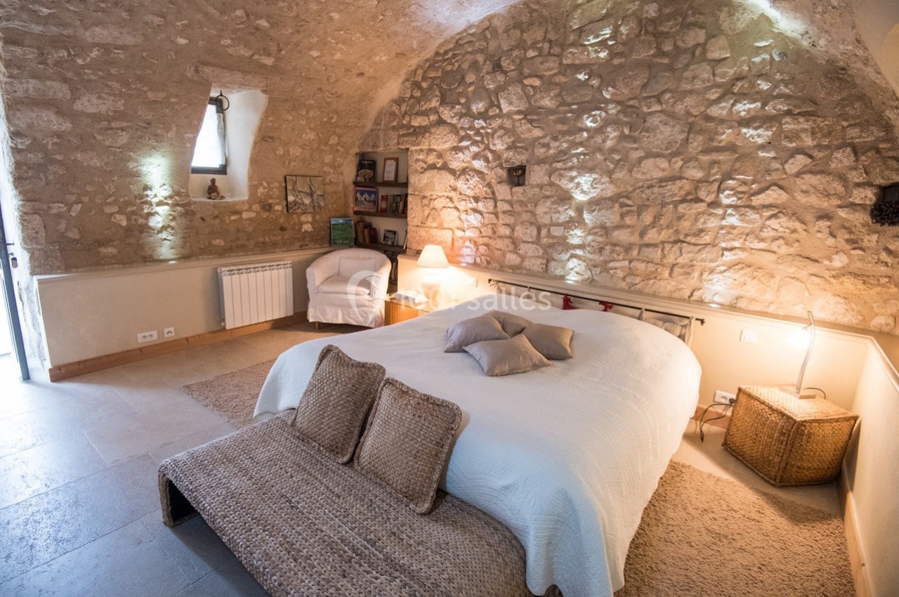 Chambre avec murs en pierre apparente, lit double, éclairage doux et mobilier en osier dans un cadre rustique.