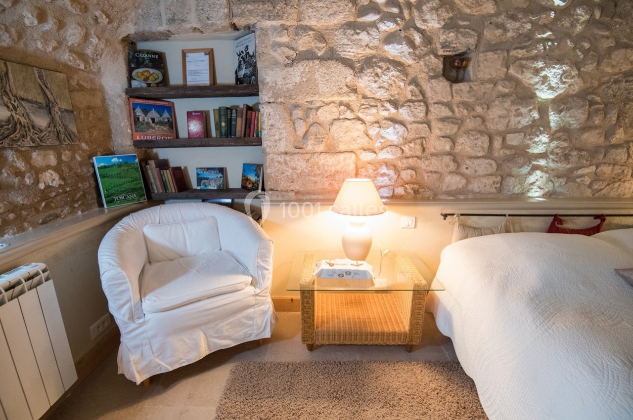 Fauteuil blanc, table basse en osier, lampe allumée et étagère avec livres dans une chambre aux murs en pierre.