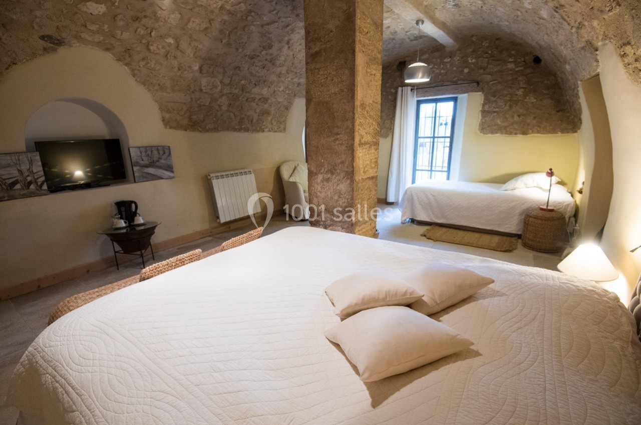 Chambre en pierre voûtée avec deux lits, une télévision murale, une fenêtre et un éclairage chaleureux.