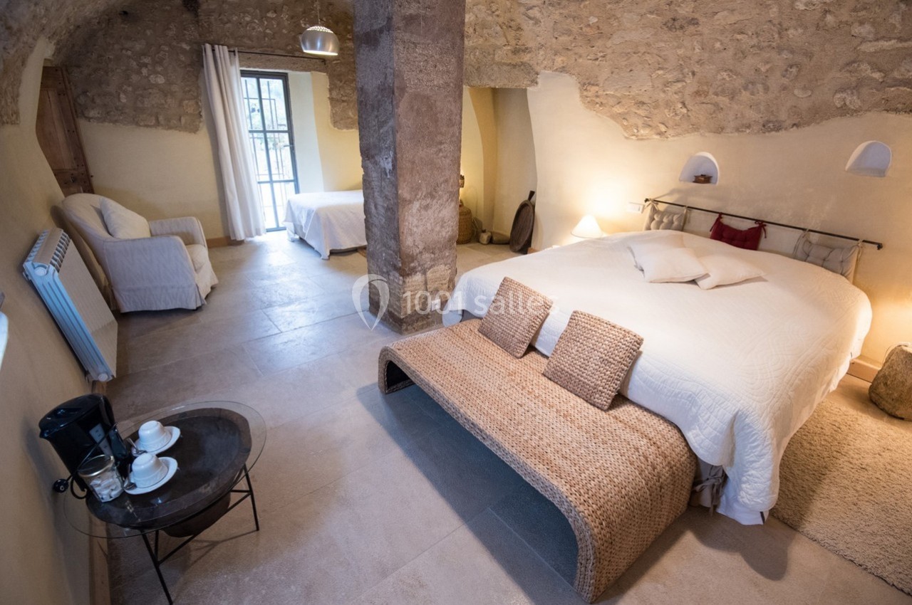 Chambre en pierre avec deux lits, fauteuil, table basse et lumière naturelle entrant par une porte-fenêtre.