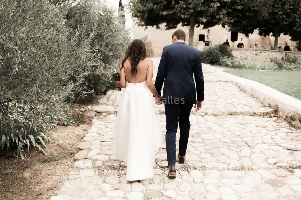 Un couple habillé élégamment marche main dans la main sur un chemin pavé bordé de verdure.