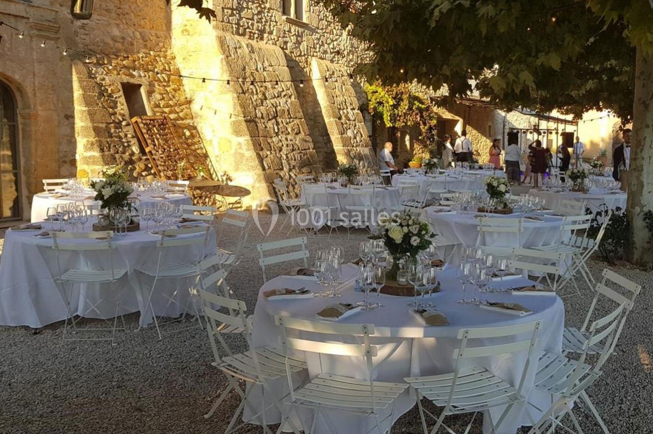 Tables rondes décorées pour un événement en extérieur, disposées sur un sol en gravier près d'un bâtiment en pierre.
