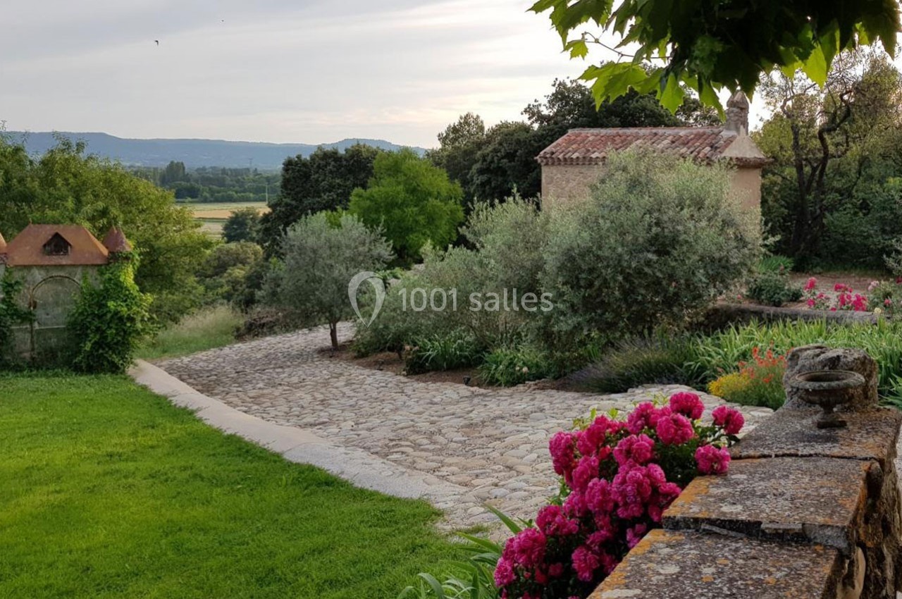 Allée pavée bordée de fleurs et d'arbustes, menant à une petite maison en pierre dans un cadre champêtre.
