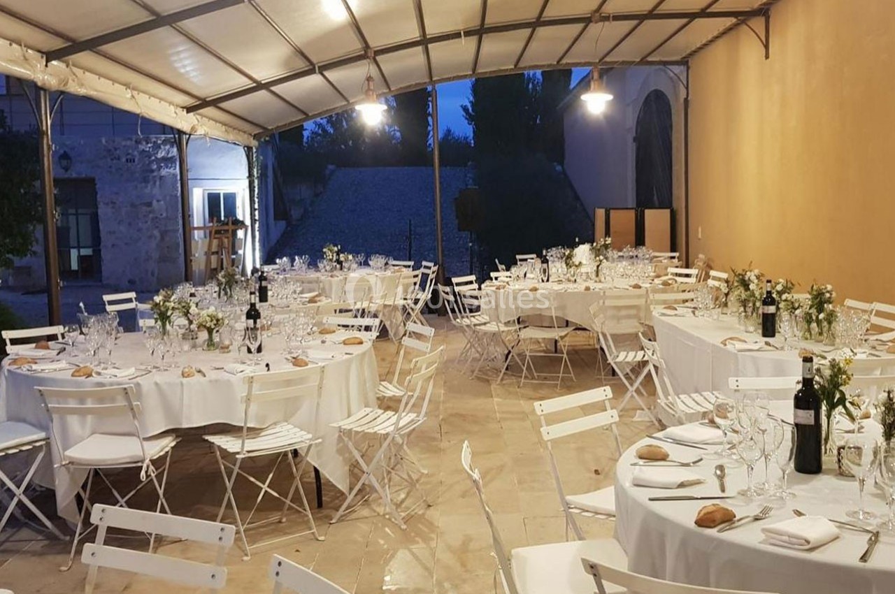 Salle couverte aménagée pour un dîner, avec des tables rondes dressées, chaises blanches et décorations florales.